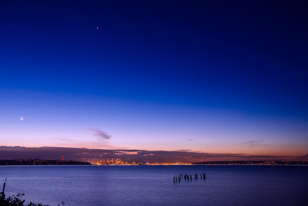 Nighttime view of Seattle city skyline across calm water under a twilight sky with moon and stars visible, USA.