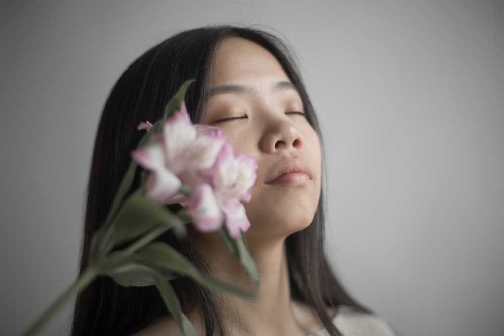 Woman with closed eyes holding a pink and white flower with green leaves close to her face in a neutral studio setting, Art, portrait, emotional.