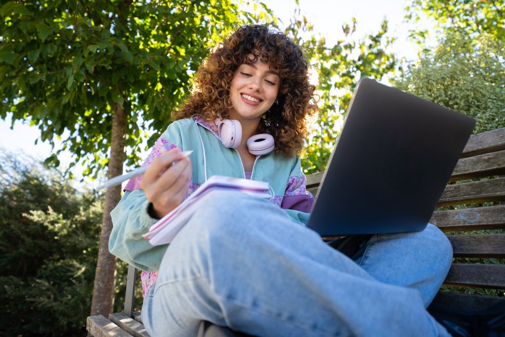 Woman sits on a wooden bench in a park writing in a notebook while using a laptop outdoors under daylight, Spain.