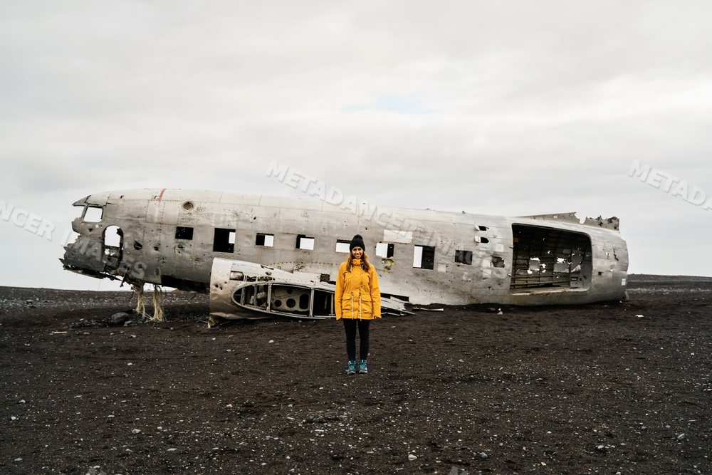 A white woman stands before an airplane wreck on black volcanic soil