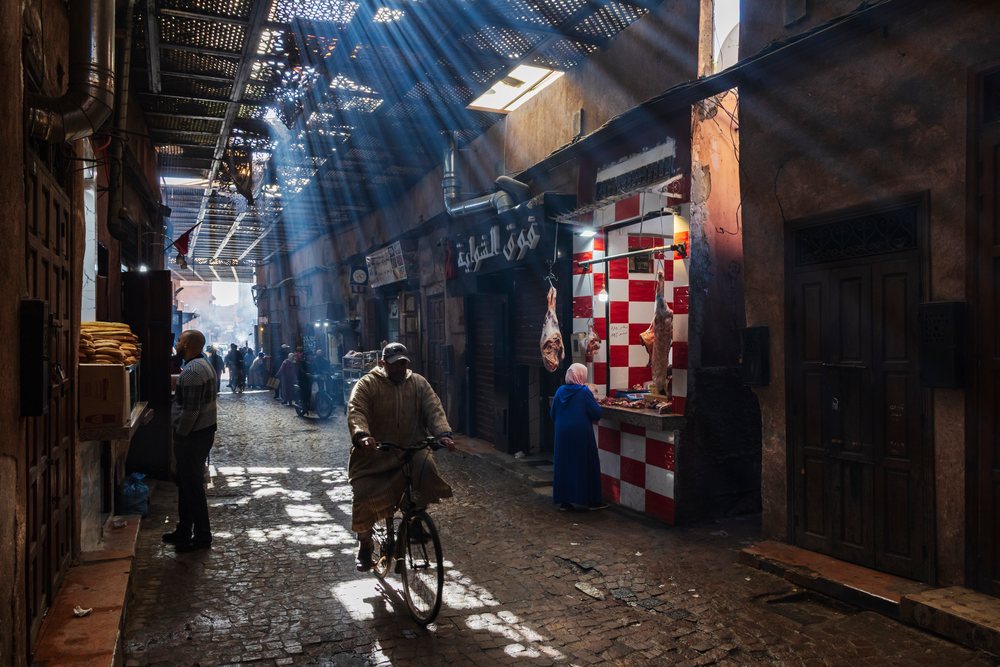 Light Rays, sunlight streams through the souq roof illuminating a bustling Marrakech market street with shoppers and a man cycling, Lifestyle, Landscape, Culture, Marrakesh-Safi Region, Morocco.