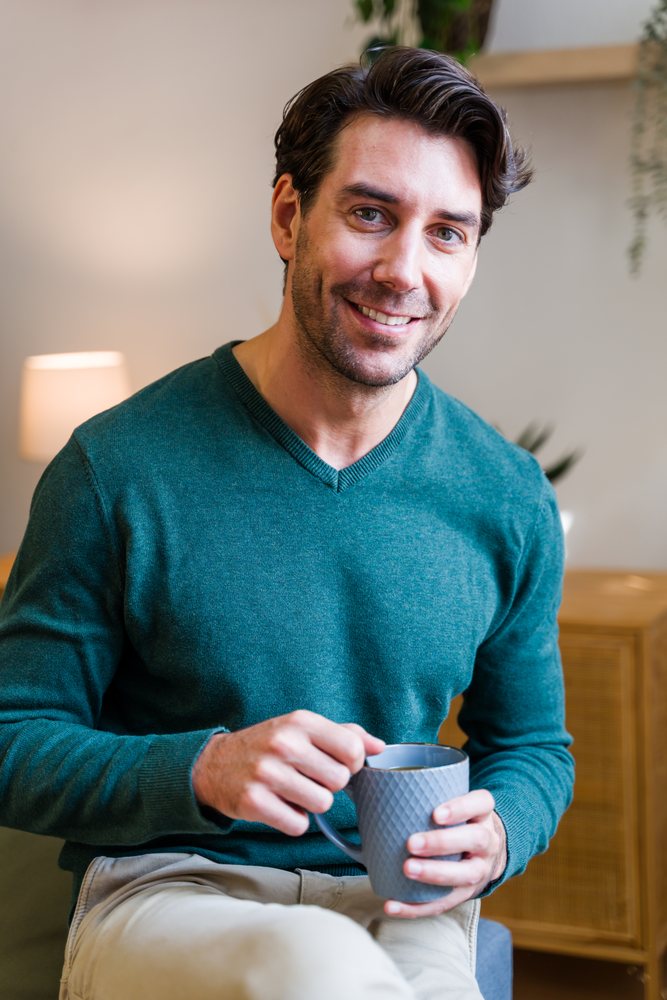 Man smiles enjoying coffee indoors while seated in a cozy living room with lamp and plants, Spain.