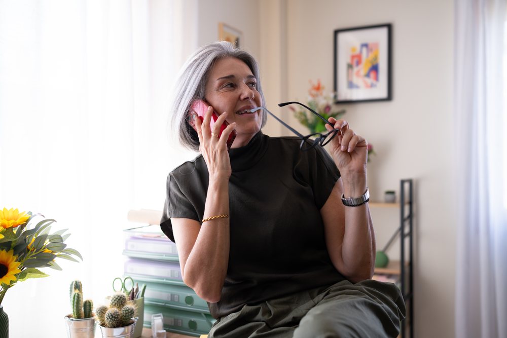 Woman with grey hair holding glasses and phone in a home office with plants and office supplies, Spain.