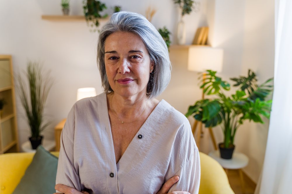Older woman with gray hair sitting on a yellow sofa in a softly lit living room interior, Spain.