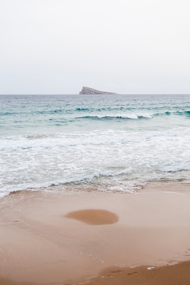 Ocean waves rolling onto a sandy beach with a rocky island visible in the Philippines under a clear sky.