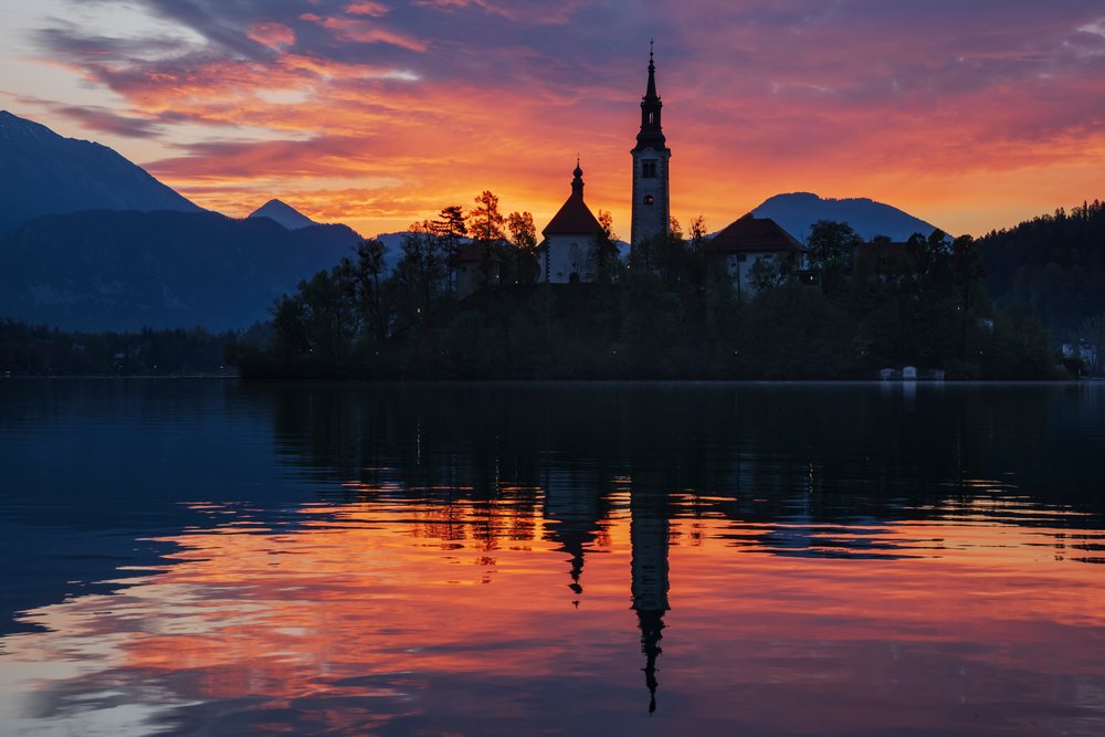 Silhouetted Church of the Assumption of the Virgin Mary on an island at Lake Bled during a colorful Sunrise with mountain reflection, landscape, cityscape, Slovenia.