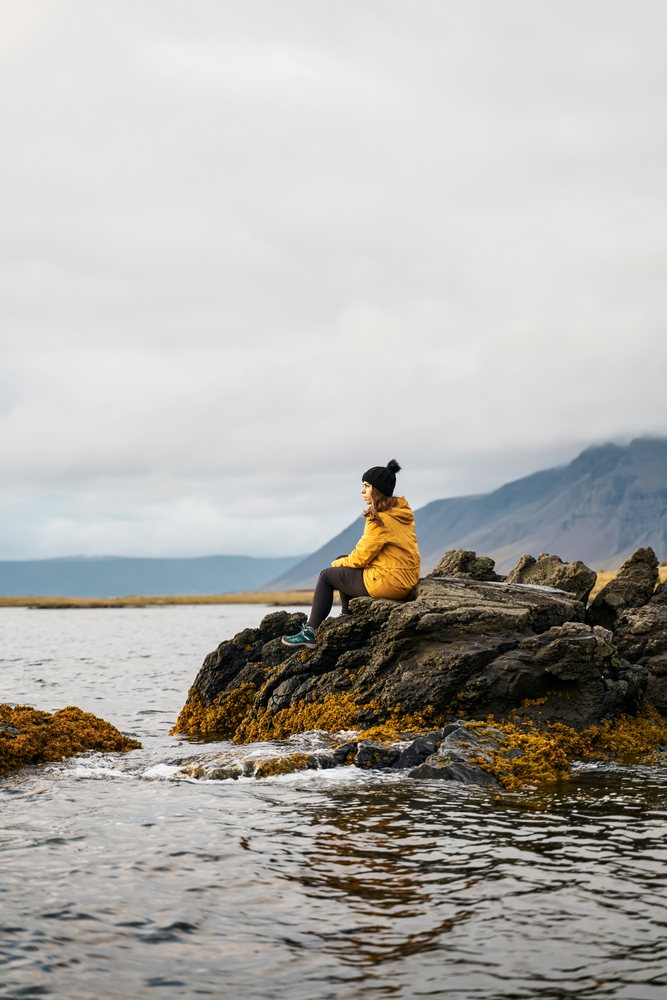A white woman sitting on rocks by the water with mountains