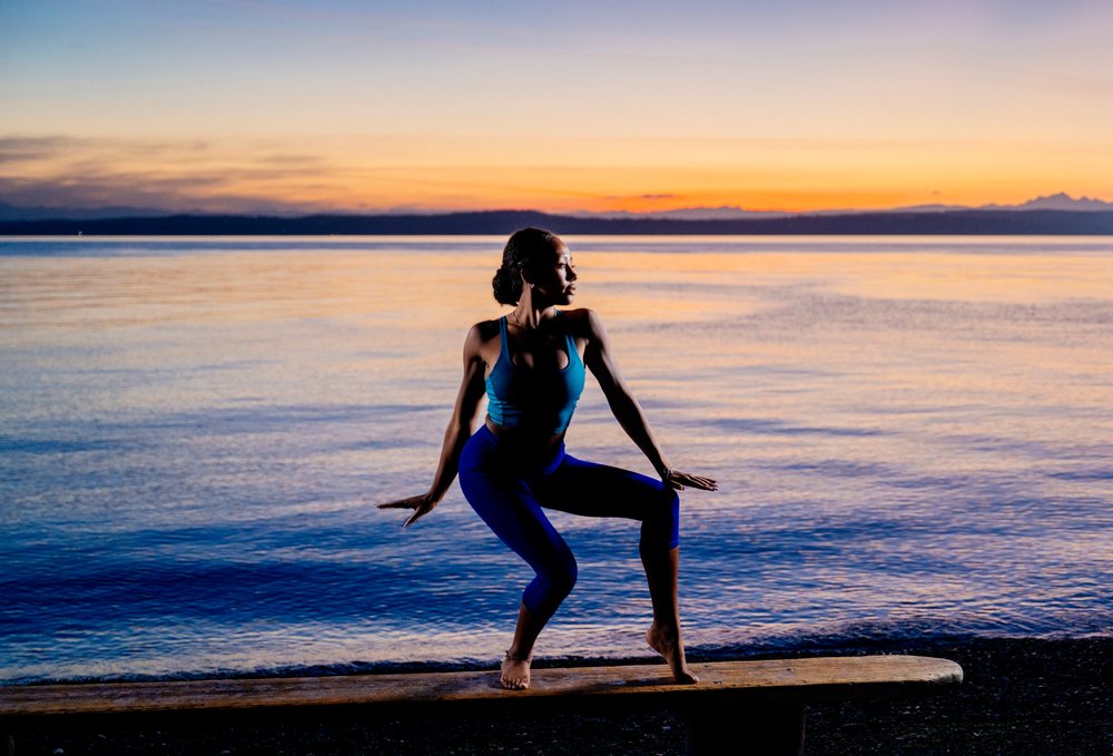 Woman practicing yoga stretching pose on a bench by the seaside at sunset, USA.