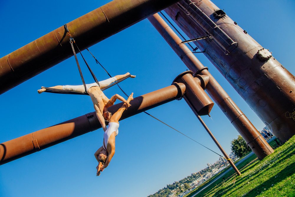 Two aerialists perform acrobatic moves on aerial silks attached to industrial pipes outdoors on a clear blue day, Seattle, WA, USA.