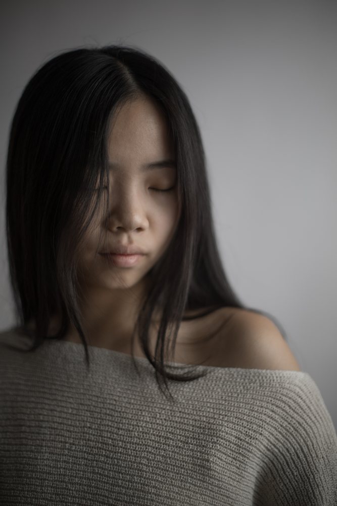 Portrait of an Asian woman with closed eyes wearing a gray sweater with an exposed shoulder in a studio setting, Art, emotional.