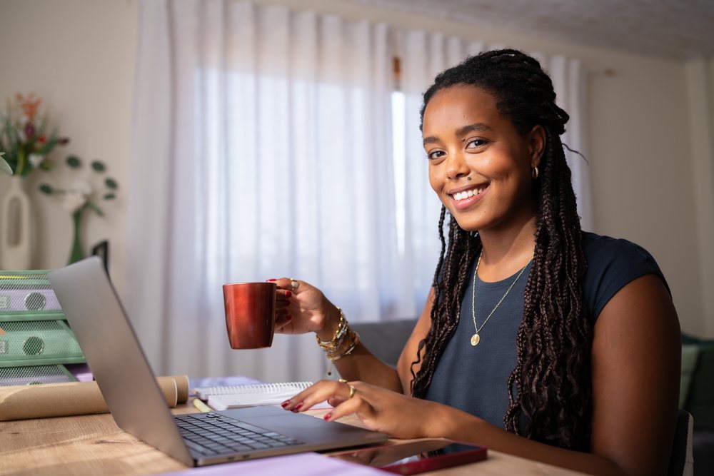 A young woman with dreadlocks is smiling while typing on her laptop and holding a cup of coffee in a home office, remote work, student, Spain.