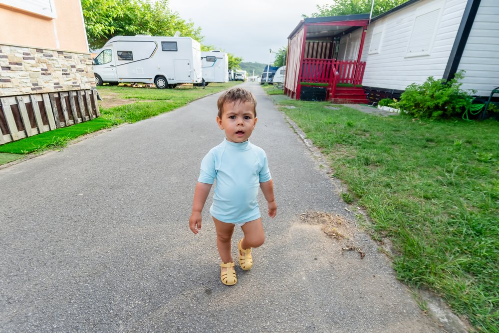 Toddler boy walking on a paved path at a campground with white campervans parked on grass in Spain during daytime, kids.