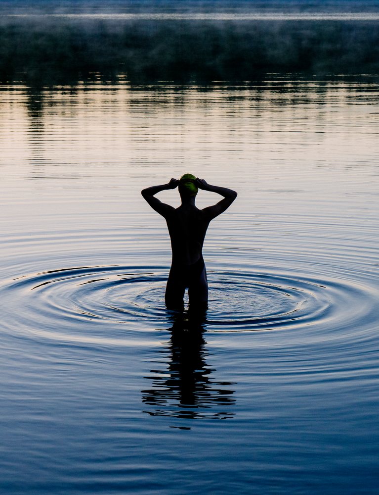 Woman wearing a swimming cap and swimsuit standing in calm lake water creating ripples at sunset, USA.