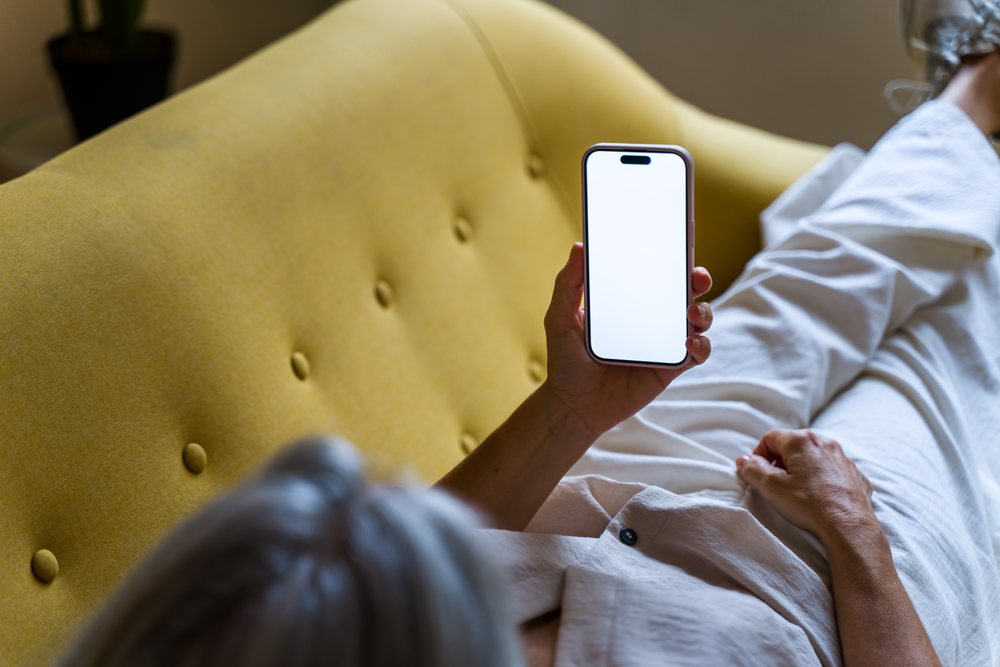 Person reclining indoors on a yellow sofa holding a smartphone with a blank screen, Spain.