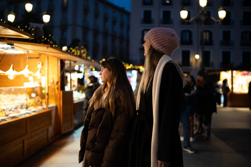 Two women browse illuminated Christmas market stalls at night during the holiday season, Madrid, Spain.