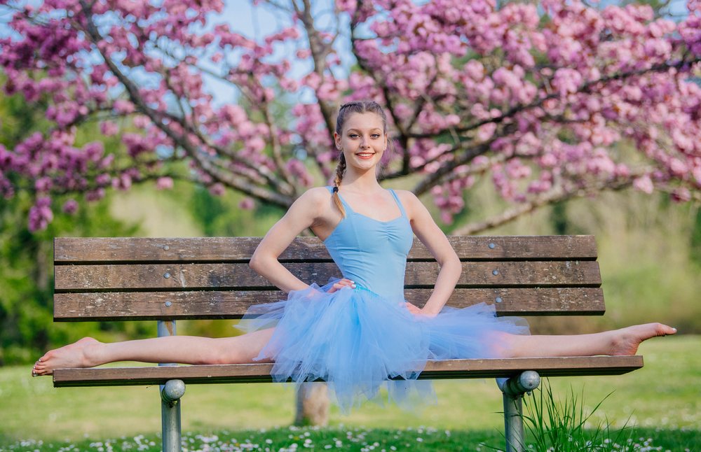 Ballet dancer in a blue tutu performs a split on a wooden bench outdoors with pink flowering trees, USA.