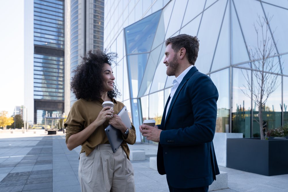 Two business people holding coffee cups and engaging in conversation outside a glass office building in an urban downtown area during daytime, Madrid, Spain.
