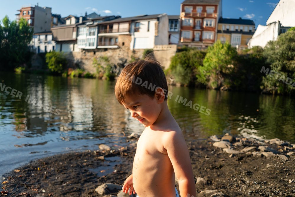 Shirtless white boy stands by the riverbank with buildings