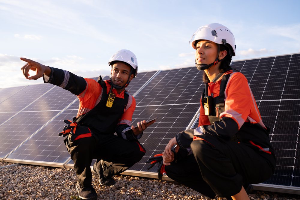 Two technicians wearing white helmets and orange and black workwear inspect solar panels mounted on gravel outdoors in sunlight, Madrid, Spain.