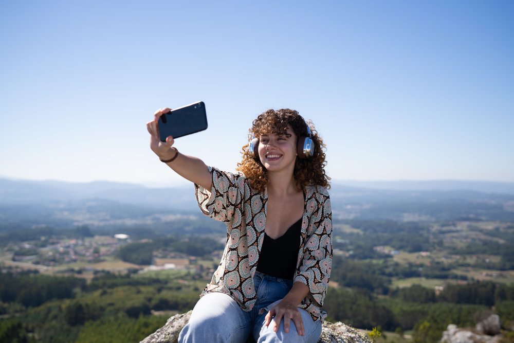 Woman sitting on a hilltop wearing headphones and taking a selfie with a smartphone overlooking a scenic forest landscape, Pico Sacro, Galicia, Spain.