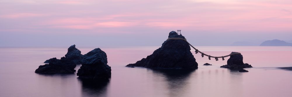 Rocks connected by a shimenawa rope and topped by a torii gate in calm ocean waters at dawn.