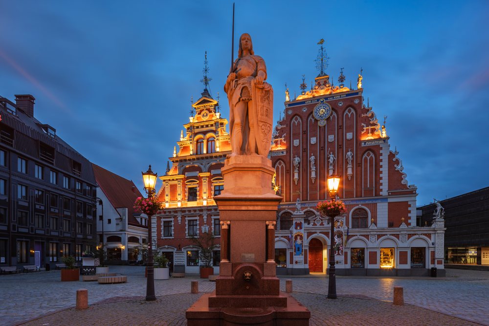 The illuminated House of the Black Heads and the large stone statue of Roland stand prominently in Riga’s historic Town Hall Square at dusk, cityscape, Latvia, Baltic States.