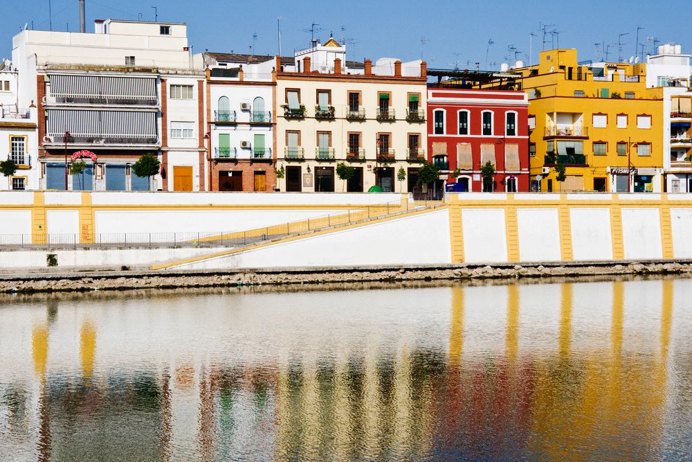 Colorful multi-story buildings with balconies line the Guadalquivir riverfront, reflecting on the water with trees and a walkway alongside, Andalusia, Spain.