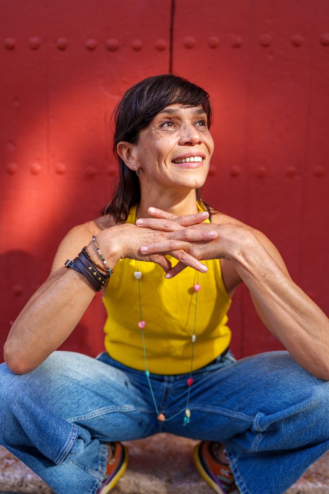 Woman wearing a yellow top and blue jeans sits smiling in front of a red wall, retro, vintage, Málaga, Spain.