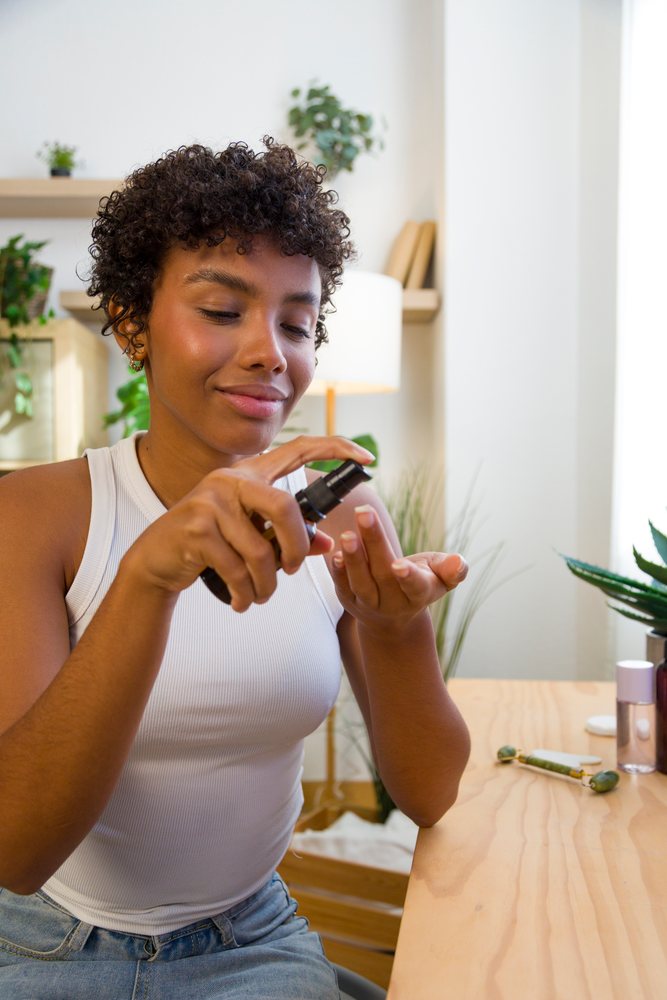 Young woman applying lotion on her hand while sitting at a wooden table at home with skincare tools nearby.
