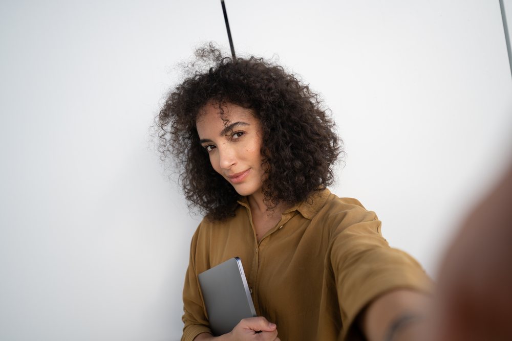 Woman with curly hair wearing a brown shirt holding a tablet and taking a selfie against a white wall indoors, Madrid, Spain.