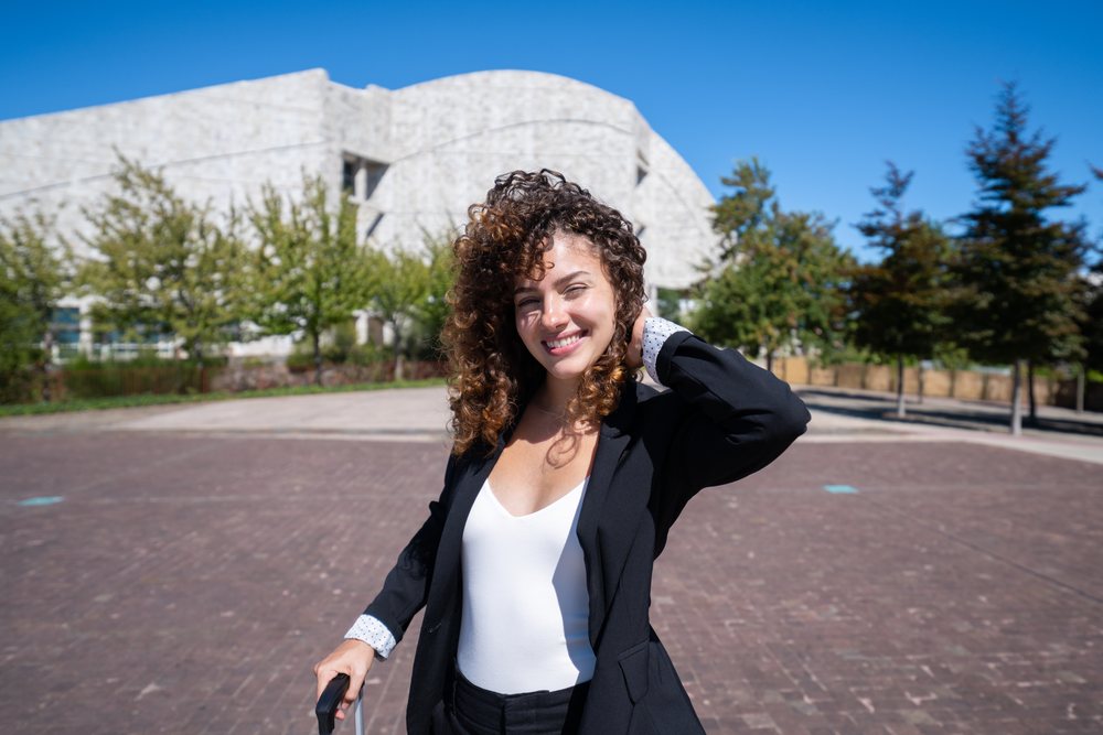Businesswoman with curly hair poses outdoors in a paved plaza near a modern building with trees in Santiago de Compostela, traveling executive, Spain, Antiago De Compostela.