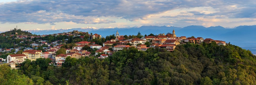Town of Sighnaghi on a hillside with red-tiled rooftops and dense forest, with the Caucasus Mountains under an early morning Sunrise sky, Georgia.
