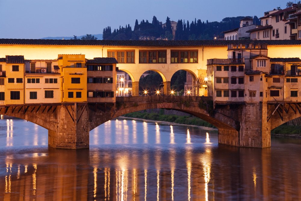Ponte Vecchio stone bridge with arches over River Arno in Florence reflecting evening lights with illuminated historic buildings and hills, Tuscany, Italy.