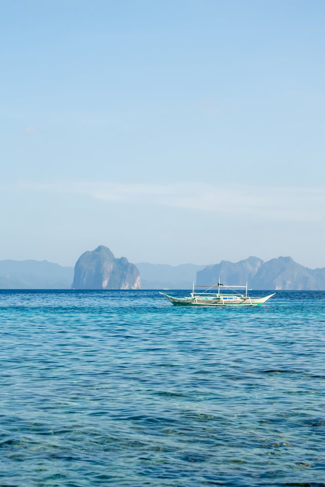 Boat floating on clear blue sea near islands under a blue sky with sparse clouds, Philippines.