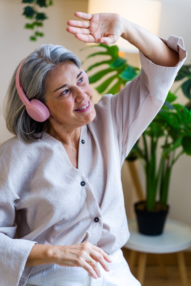 Middle-aged woman with gray hair wearing pink headphones smiles and dances indoors near a houseplant, Spain.