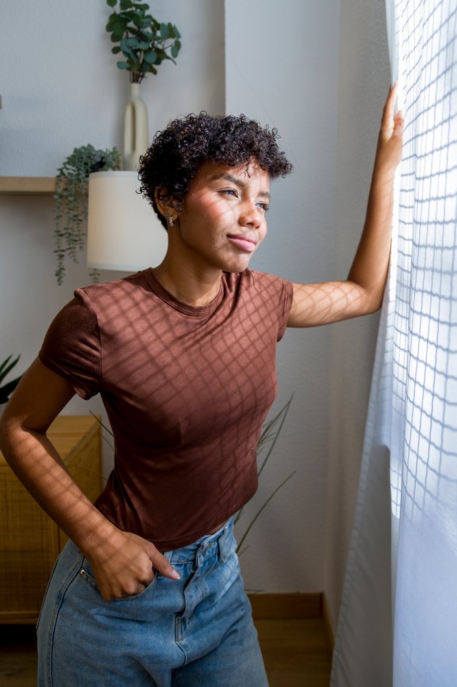 Young woman in casual clothing stands indoors looking thoughtfully out a window with white curtains at home.