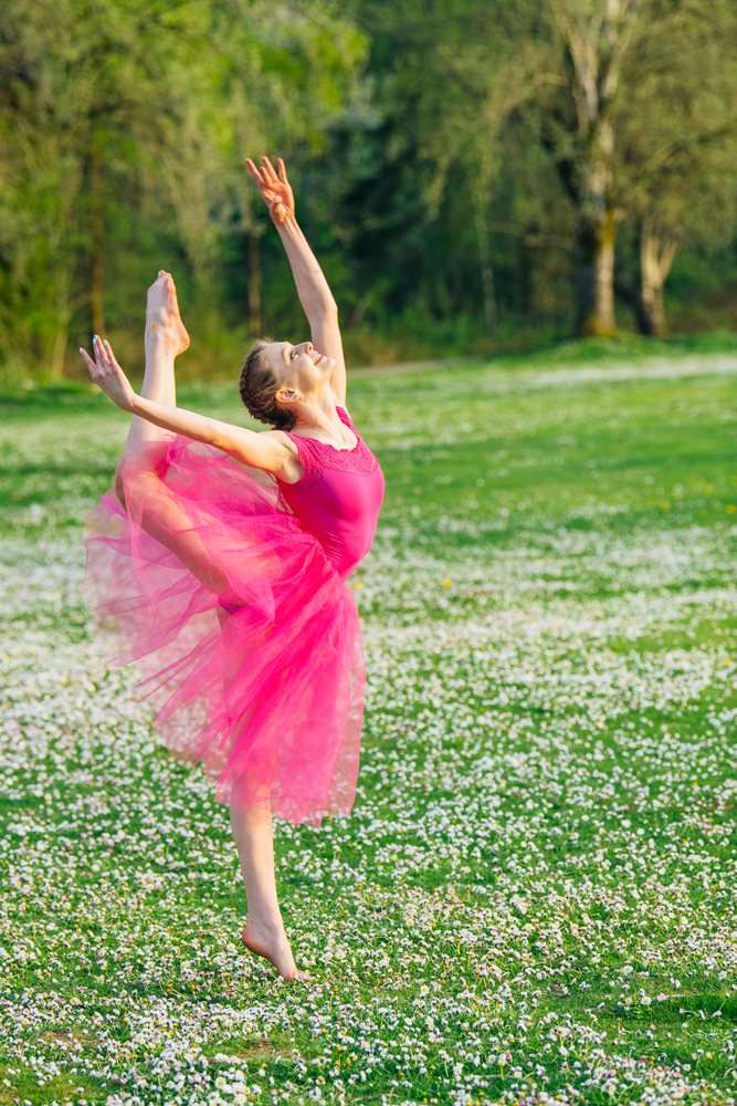 Ballet dancer in a pink tutu poses gracefully outdoors in a grassy meadow with white flowers and trees, USA.