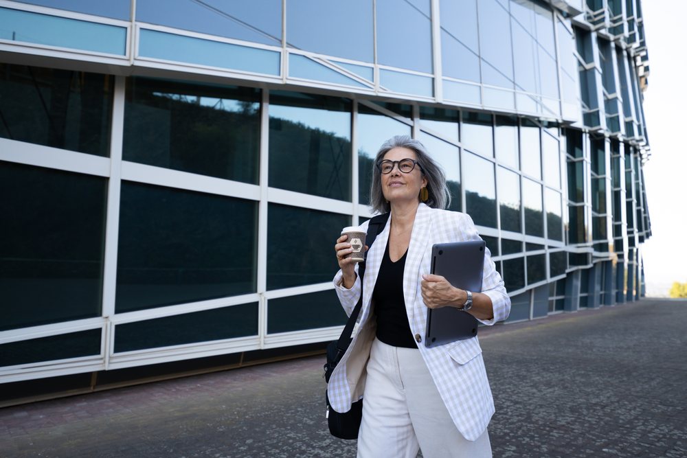 Mature businesswoman with gray hair and glasses walks on a sidewalk outside a modern glass building, holding a takeaway coffee cup and carrying a tablet, businesswomen, Spain.