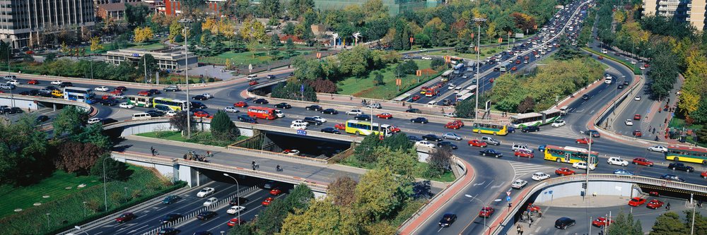 Multiple road overpasses and busy urban roads with cars, buses, and pedestrians near a green park area.