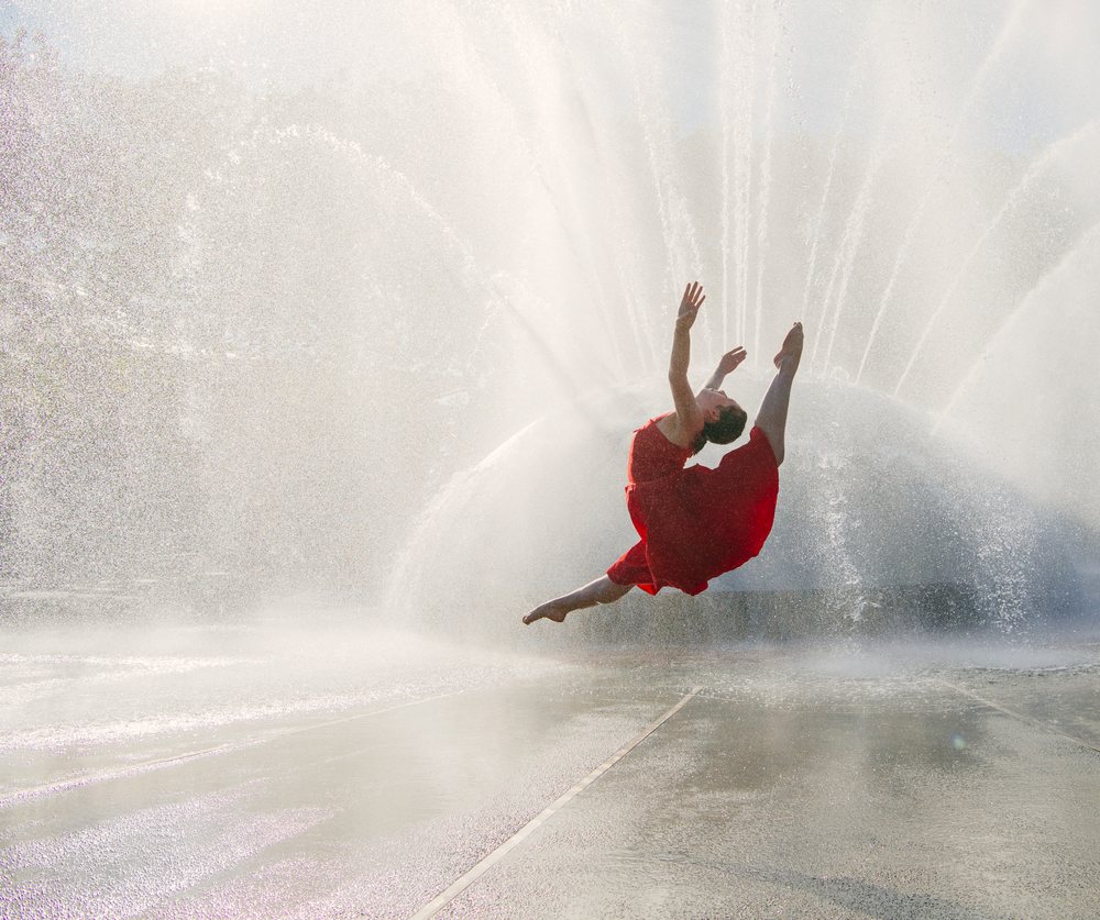 Female dancer leaps gracefully outdoors in front of a spraying water fountain on concrete ground in sunlight, USA.