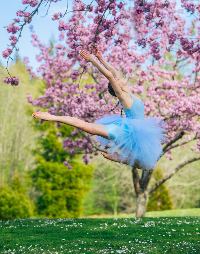 Woman ballet dancer wearing a blue tutu leaps gracefully outdoors near a flowering tree with pink blossoms in spring in a USA park.