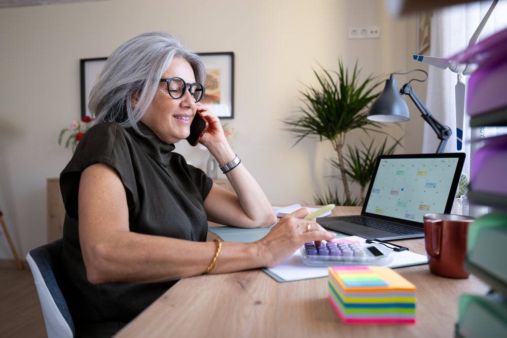 Woman smiling while talking on phone and using calculator at a home office desk with laptop and calendar, Spain.