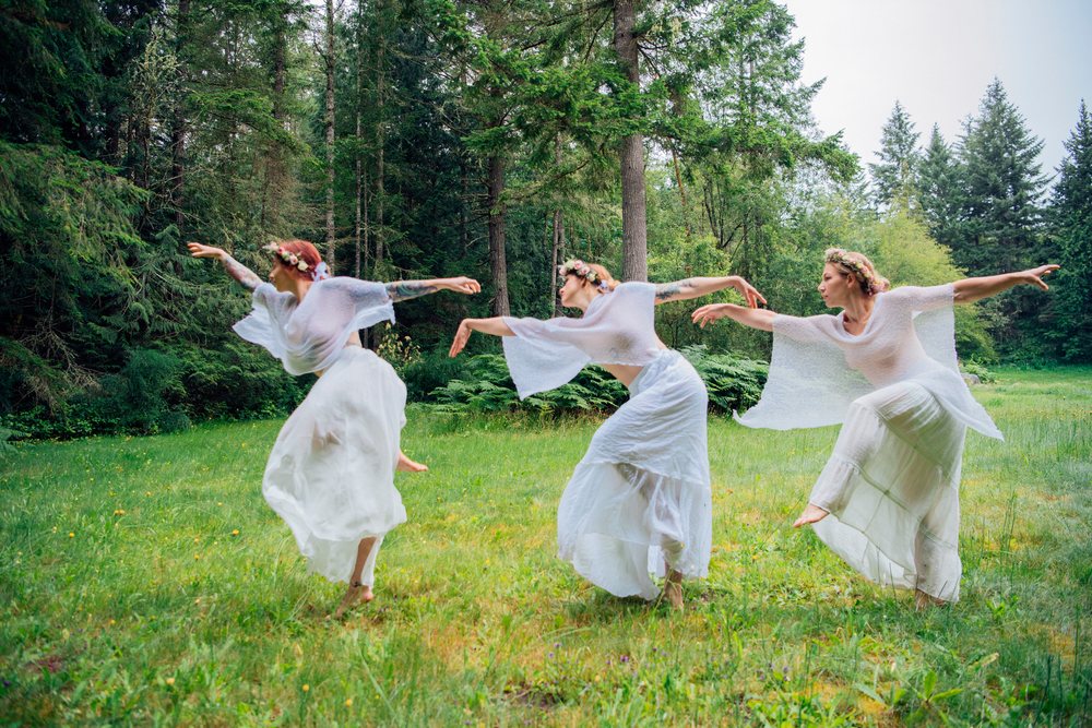 Three women dancing barefoot in white dresses with flower crowns in a forest meadow, muses, USA.