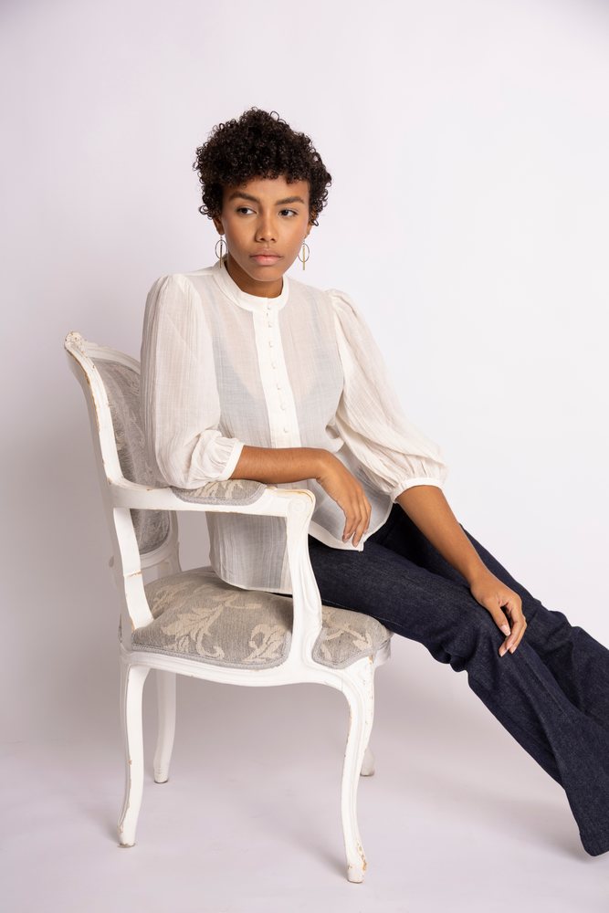Woman sits in a white vintage-style chair wearing a puffed sleeve white blouse and dark pants against a plain background, fashion, young culture.