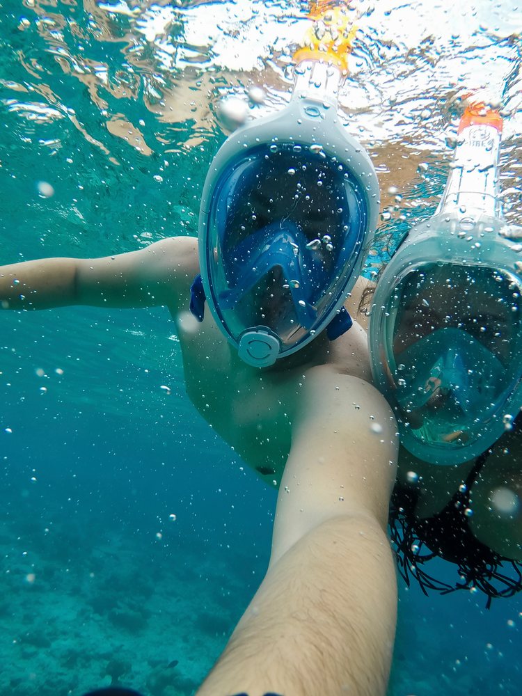Two people wearing full-face snorkel masks swimming underwater near a coral reef in the, Maldives.