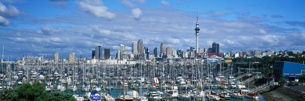 Numerous boats with sailboat masts docked in Auckland marina with urban skyline and Sky Tower under a partly cloudy sky, Marina Auckland, New Zealand.