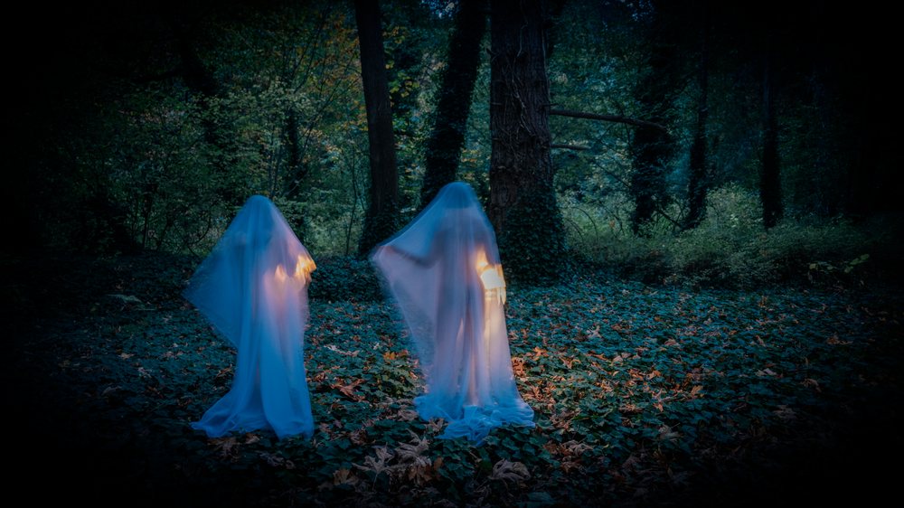 Two glowing ghost figures with candlelight stand in a dark, ivy-covered forest floor at night, ghosts, USA.