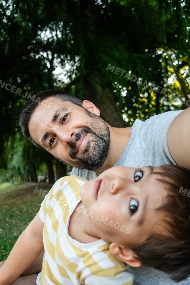 A father enjoys taking a selfie with his young son outdoors in a park