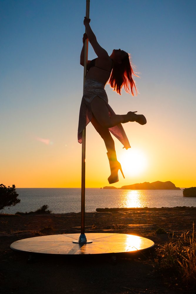 Silhouetted female pole dancer performing at sunset on a rocky shore, depicting empowerment and movement, Ibiza, Spain.