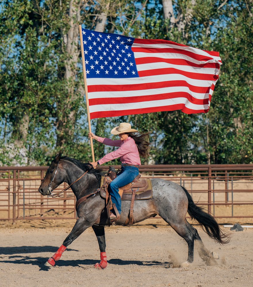 Cowgirl rides a galloping horse holding a large American flag in a dusty rodeo arena outdoors, Southwest USA.